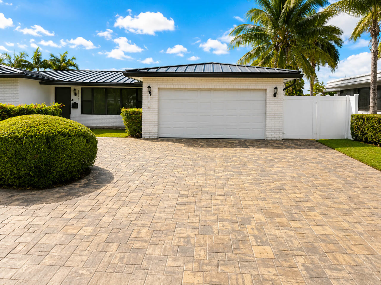 Paver driveway in a beige running bond pattern at a white brick home with a black standing seam metal roof in Coral Springs