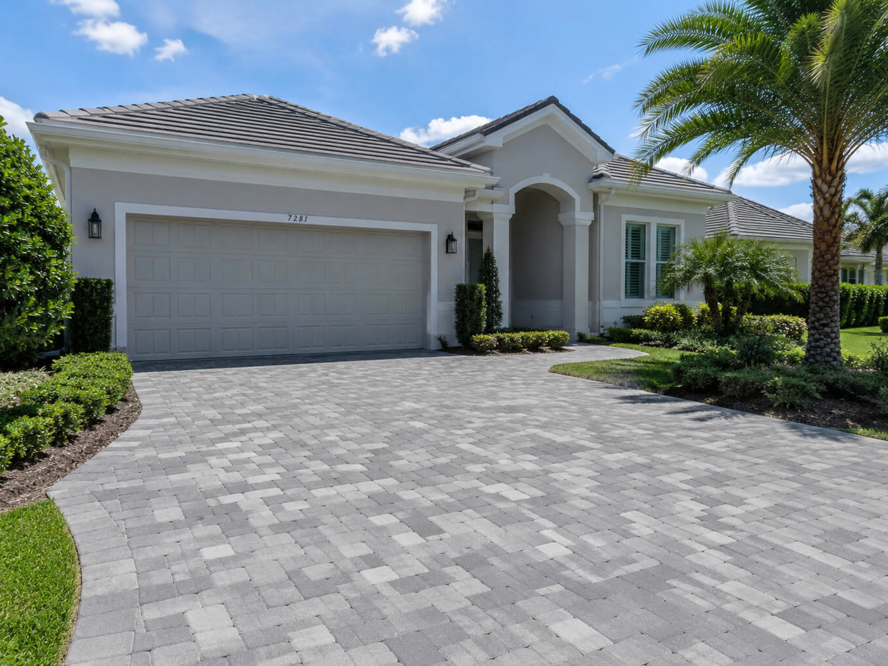 Gray tumbled paver driveway curving up to a two car garage framed by clipped hedges and palms for a Greenacres homeowner