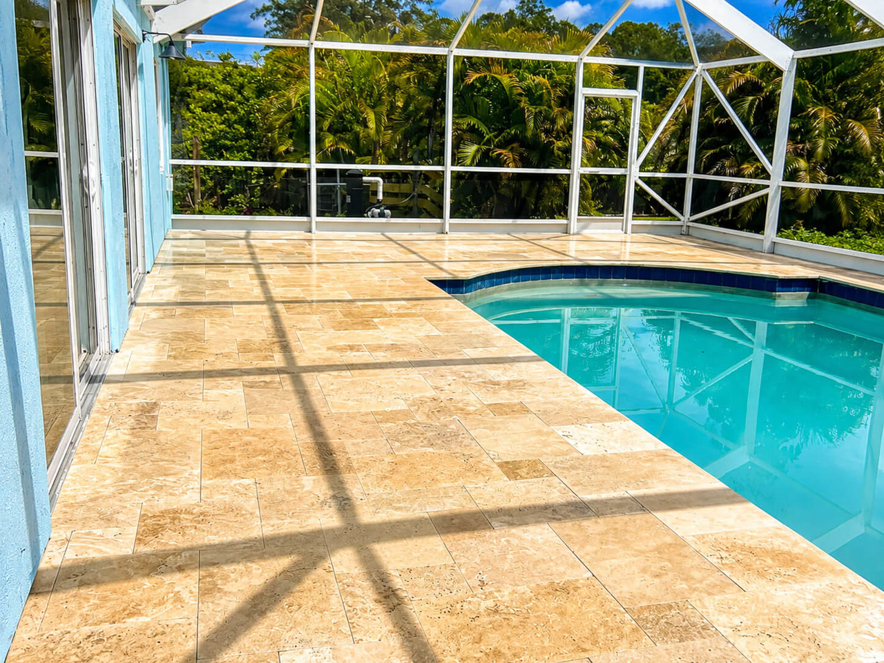 Sand toned travertine paver pool deck under a white screen enclosure at a Juno Beach residence, with dark blue tile coping along the pool