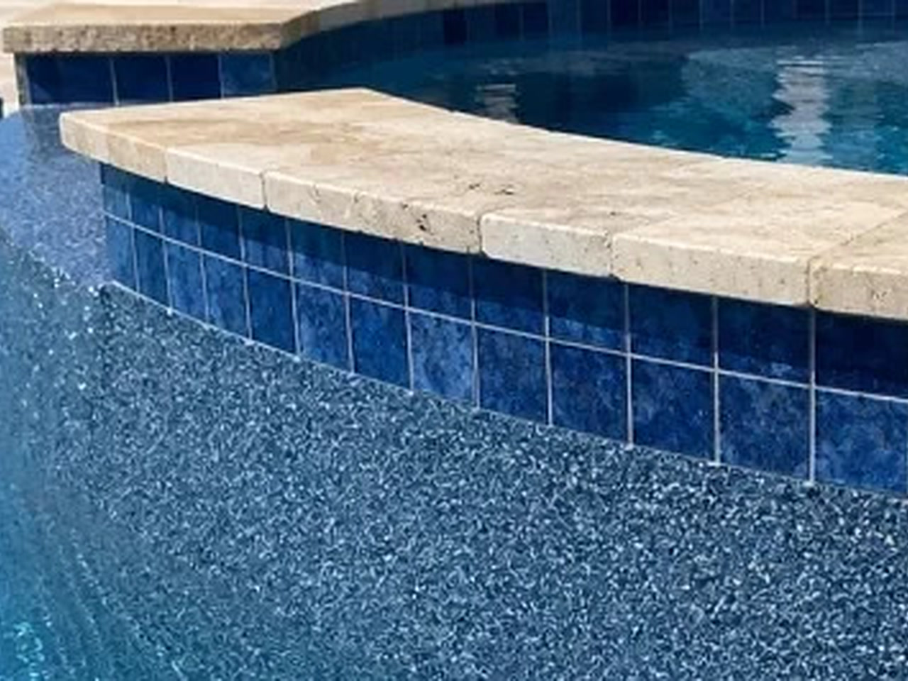 Blue pebble pool finish meeting a raised spa wall of deep blue tile capped with travertine coping on a Boca Raton property