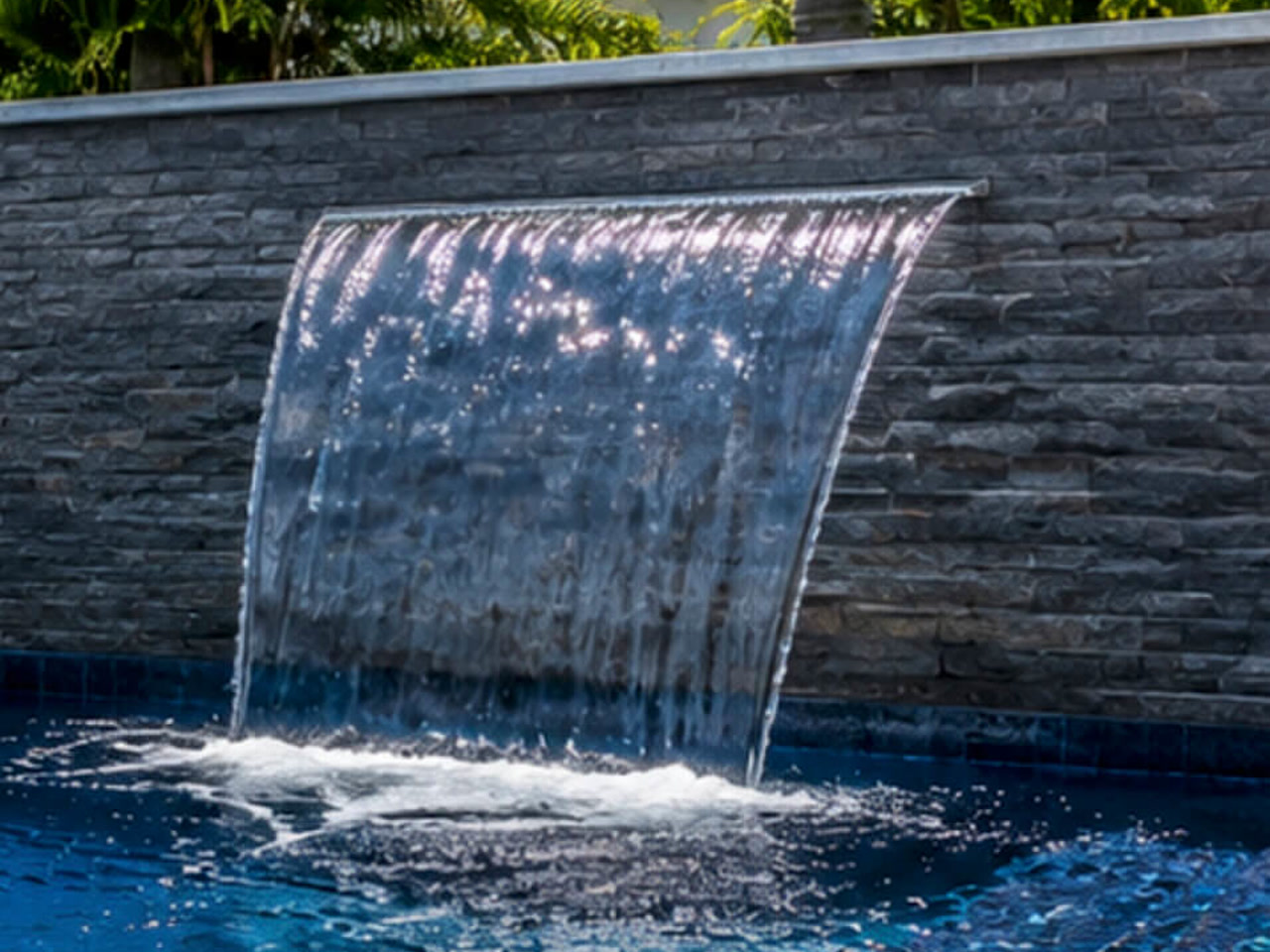 Sheer descent waterfall blade falling from a gray stacked stone wall into a deep blue pool at a Coral Springs pool