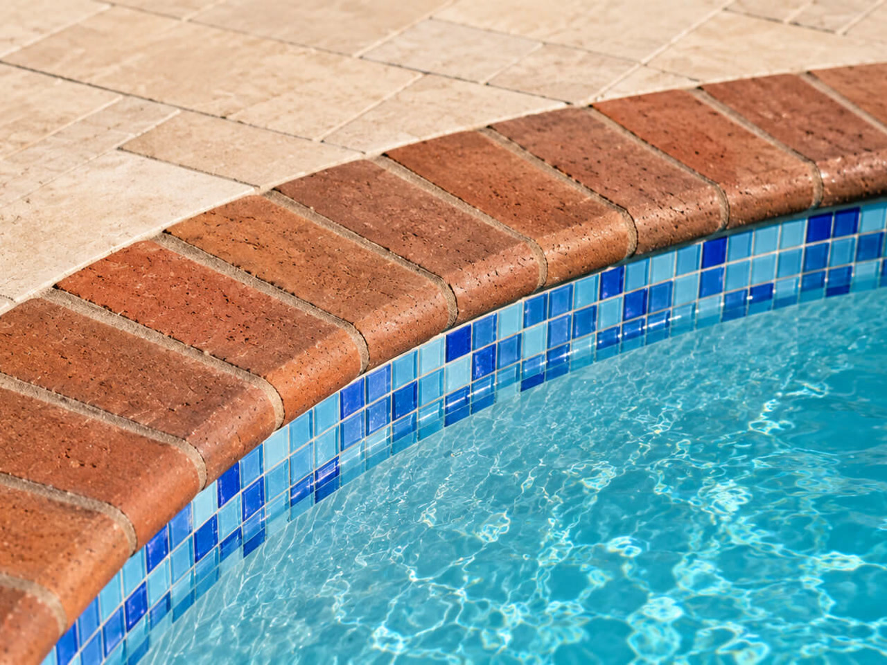Pool coping in red brick following the curve of the water over blue mosaic waterline tile, beside a travertine deck in Deerfield Beach